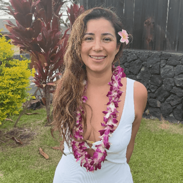 A woman with a flower lei smiles outdoors with tropical plants behind her.