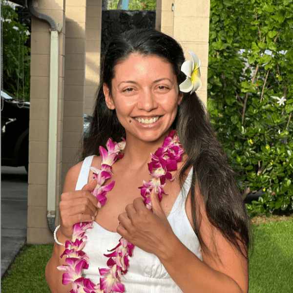 Smiling woman with flower lei and white flower in hair outdoors.