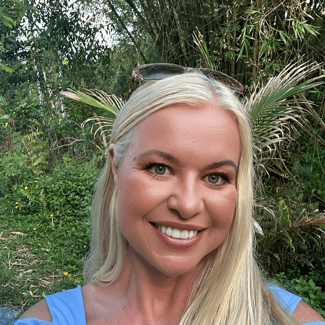 Smiling woman with long blonde hair outdoors among greenery.